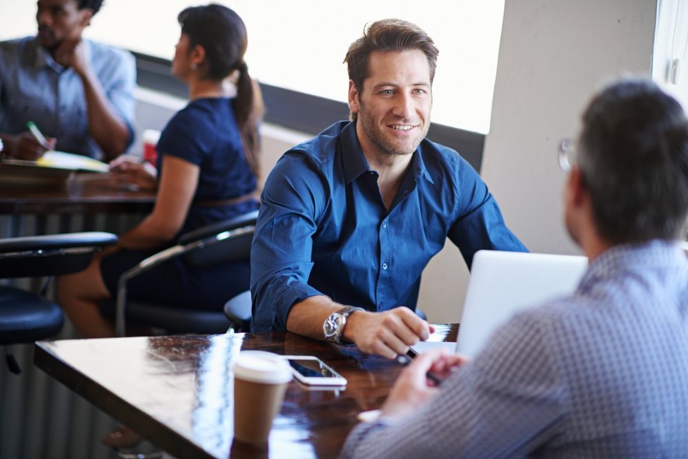 Smiling man in a blue shirt talking to another man across a desk in a busy office, with other professionals working in the background.