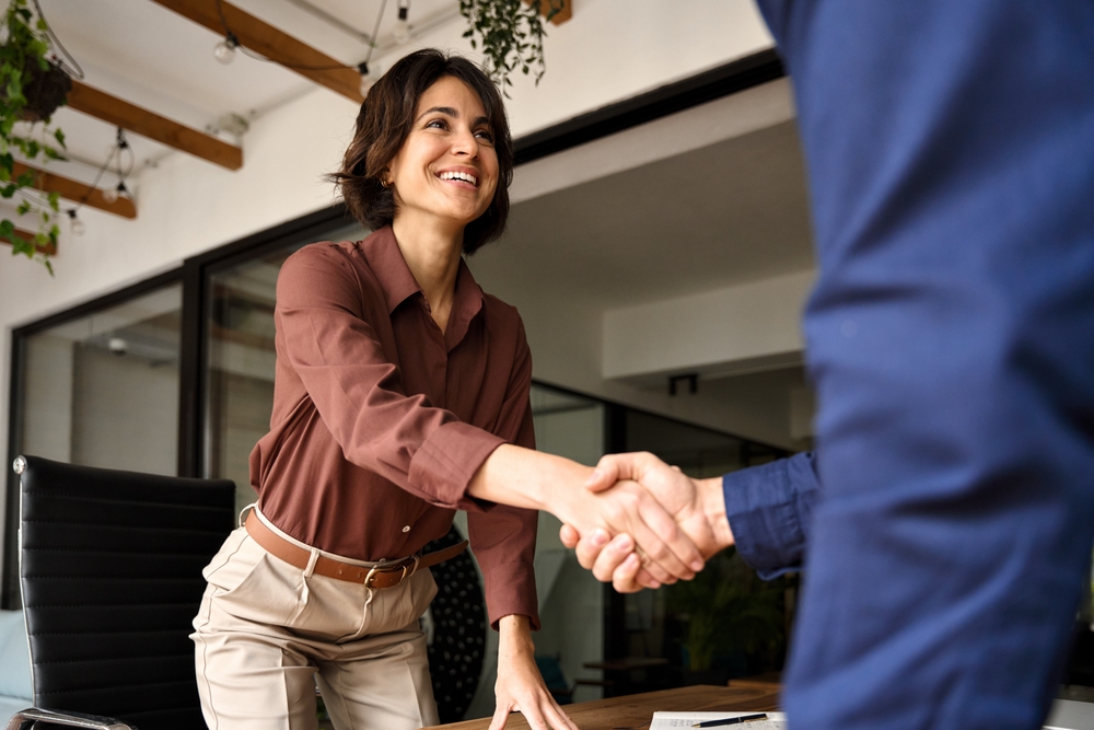 Smiling woman in business attire reaching across a desk to shake hands with a man, suggesting a positive job interview or professional agreement in a modern office