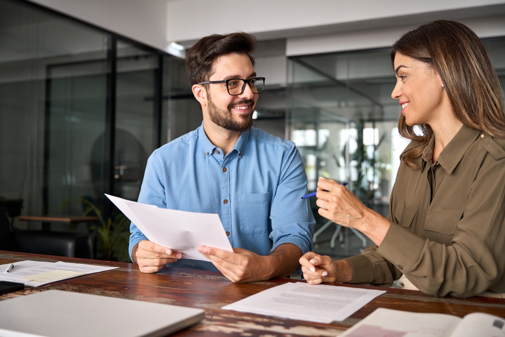 Two professionals, a man holding documents and a woman with a pen, sitting at a desk and discussing paperwork in a bright, modern office environment